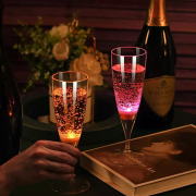 Two LED champagne glasses, yellow on the left and pink on the right illuminating in dark room with flowers, champagne, and book in the background