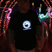 Man wearing light up t-shirt in outdoor setting with lights in the background, logo on shirt