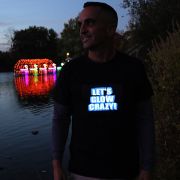 Smiling guy wearing "lets glow crazy" light up shirt outside in front of water and LED swan boats