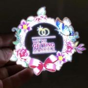 Hand holding custom EL light up panel with we're getting married text, rings, and flower wreath lighting up bright neon colors