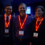 Three conference attendees wearing red LED light-up lanyards while smiling and standing together at a professional event