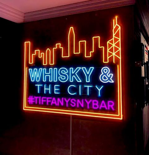 Neon sign for bar with NYC city skyline lighting up in orange and the words Whisky & The city lighting up blue with a purple tagline reading #tiffanysnybar
