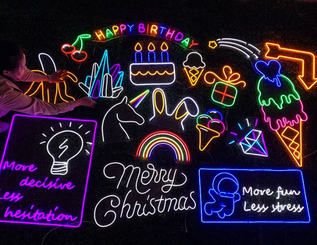 Various LED signs in a dark room lighting up bright neon colors some in specific shapes like rainbows, birthday cake, ice cream, unicorn, diamond, and others with text reading Merry Christmas, Happy birthday, More fun less stress