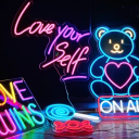 Six different neon signs in dark room with multiple colors lighting up brightly including a blue lollipop, red candy wrapper, on air, teddy bear, a rainbow sign reading "love wins" and a pink sign reading "love your self"