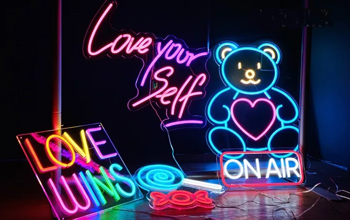 Six different neon signs in dark room with multiple colors lighting up brightly including a blue lollipop, red candy wrapper, on air, teddy bear, a rainbow sign reading "love wins" and a pink sign reading "love your self"