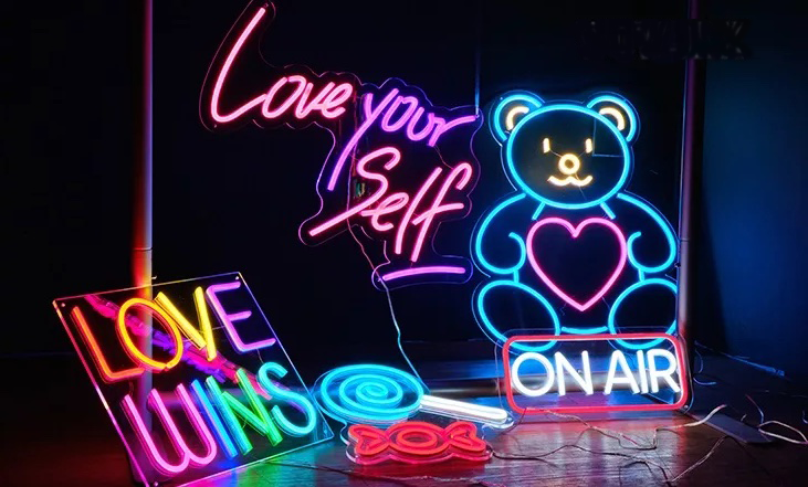 Six different neon signs in dark room with multiple colors lighting up brightly including a blue lollipop, red candy wrapper, on air, teddy bear, a rainbow sign reading "love wins" and a pink sign reading "love your self"