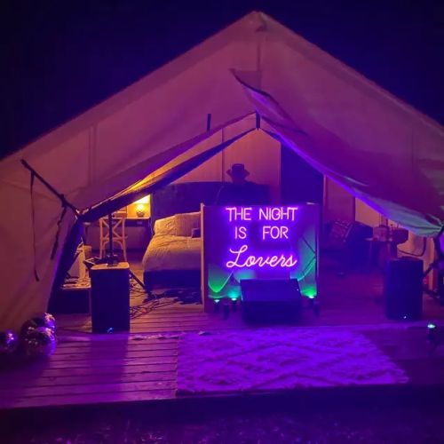 Glamping camp tent set up at night opened up to show bed in back of neon sign lighting up bright purple reading "the night is for lovers"