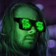 Man with long hair looking into camera while wearing animated LED sunglasses with green dollar signs glowing on them