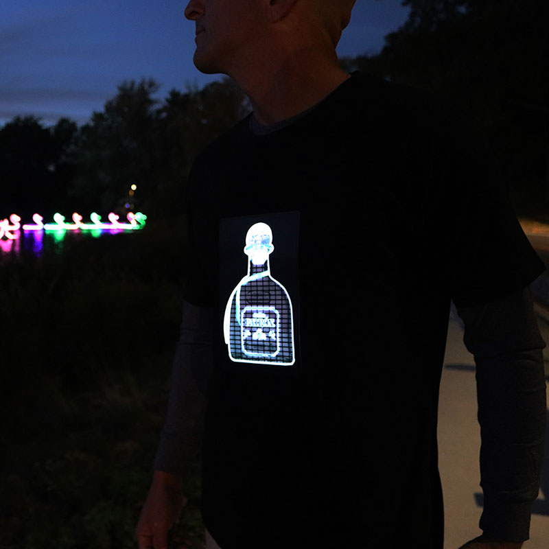 Man in dark outside wearing light up shirt with patron bottle illuminating with some neon colors lighting up in the background