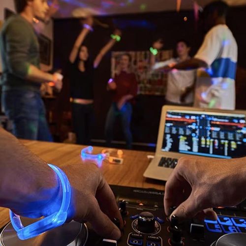 First person perspective of DJ wearing light up bracelets while operating turntable in front of crowd of dancing people wearing different colored light up bracelets