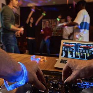 First person perspective of DJ wearing light up bracelets while operating turntable in front of crowd of dancing people wearing different colored light up bracelets